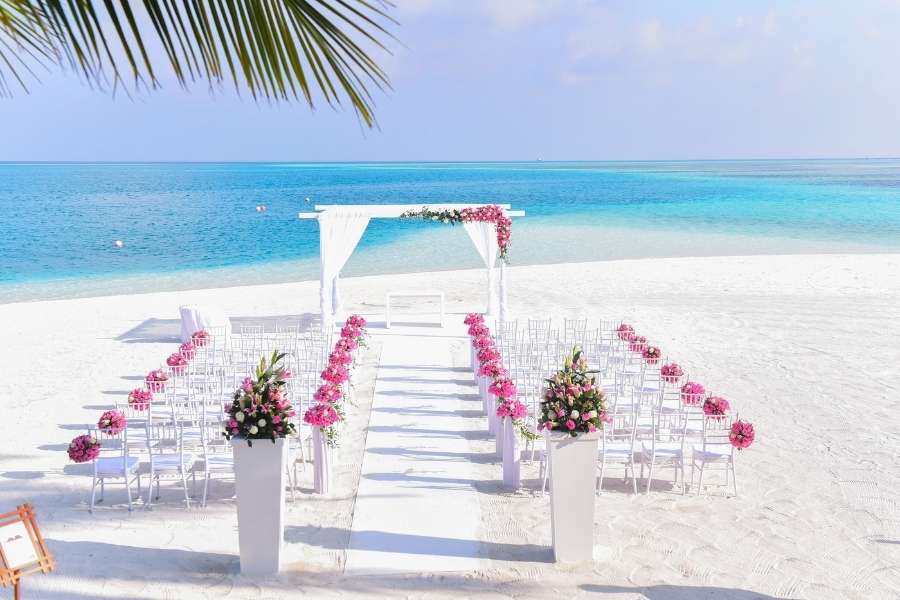Un autel de mariage décoré de fleurs fuchsia et une allée de chaises blanches sur une plage de sable fin, face à un lagon turquoise sous un ciel ensoleillé.