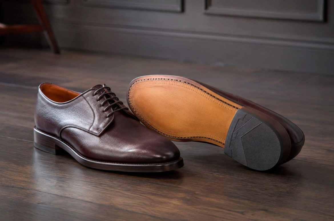 Gros plan d'une paire de chaussures Derbies en cuir marron foncé pour homme sur un parquet en bois. Une chaussure est à plat, montrant sa tige lacée, tandis que l'autre est retournée, exposant sa semelle en cuir clair cousue avec une talonnette antidérapante.